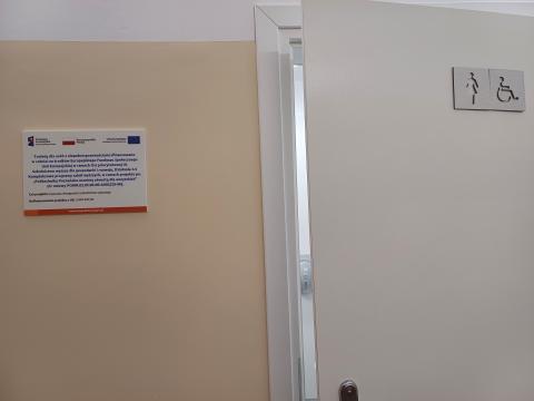Lekko uchylone drzwi do toalety z piktogramami. Tabliczka informacyjna po lewej stronie od drzwi